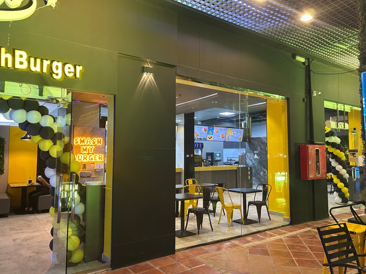 Modern burger restaurant storefront with green and yellow exterior, large windows, and black and yellow balloon decorations at mall entrance