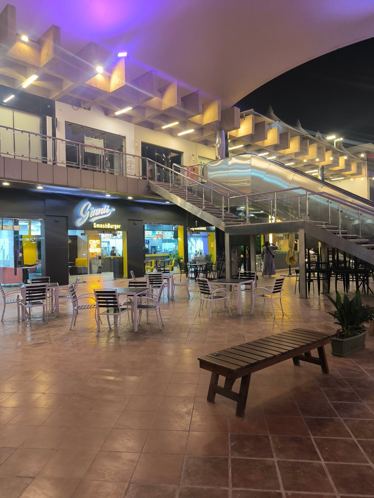 Modern indoor mall or shopping complex with dining area, illuminated storefronts, metal stairs, and contemporary architectural design at night